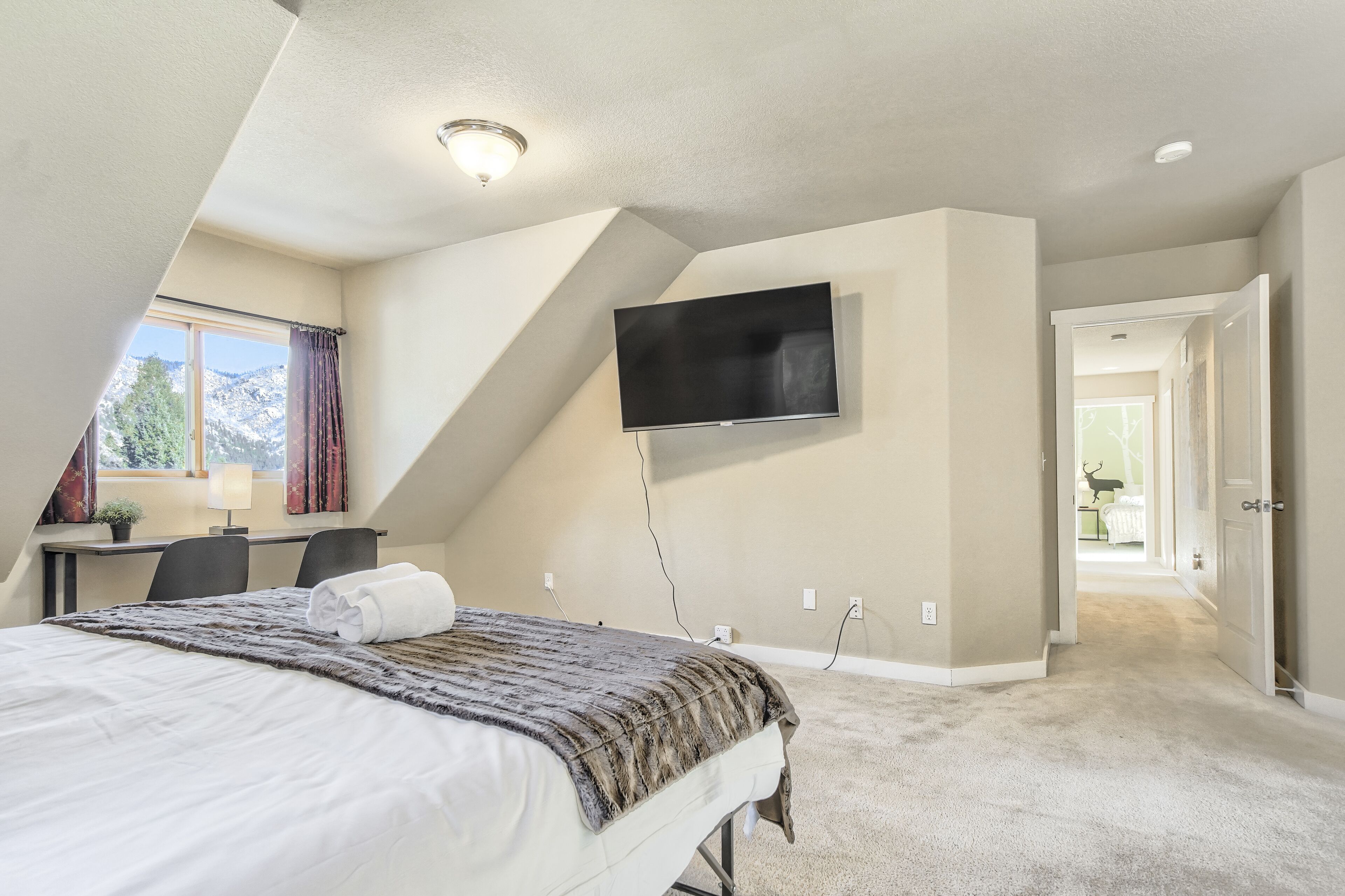 A bedroom with a bed, a desk, a TV mounted on the wall, and a view of a mountain through a window.