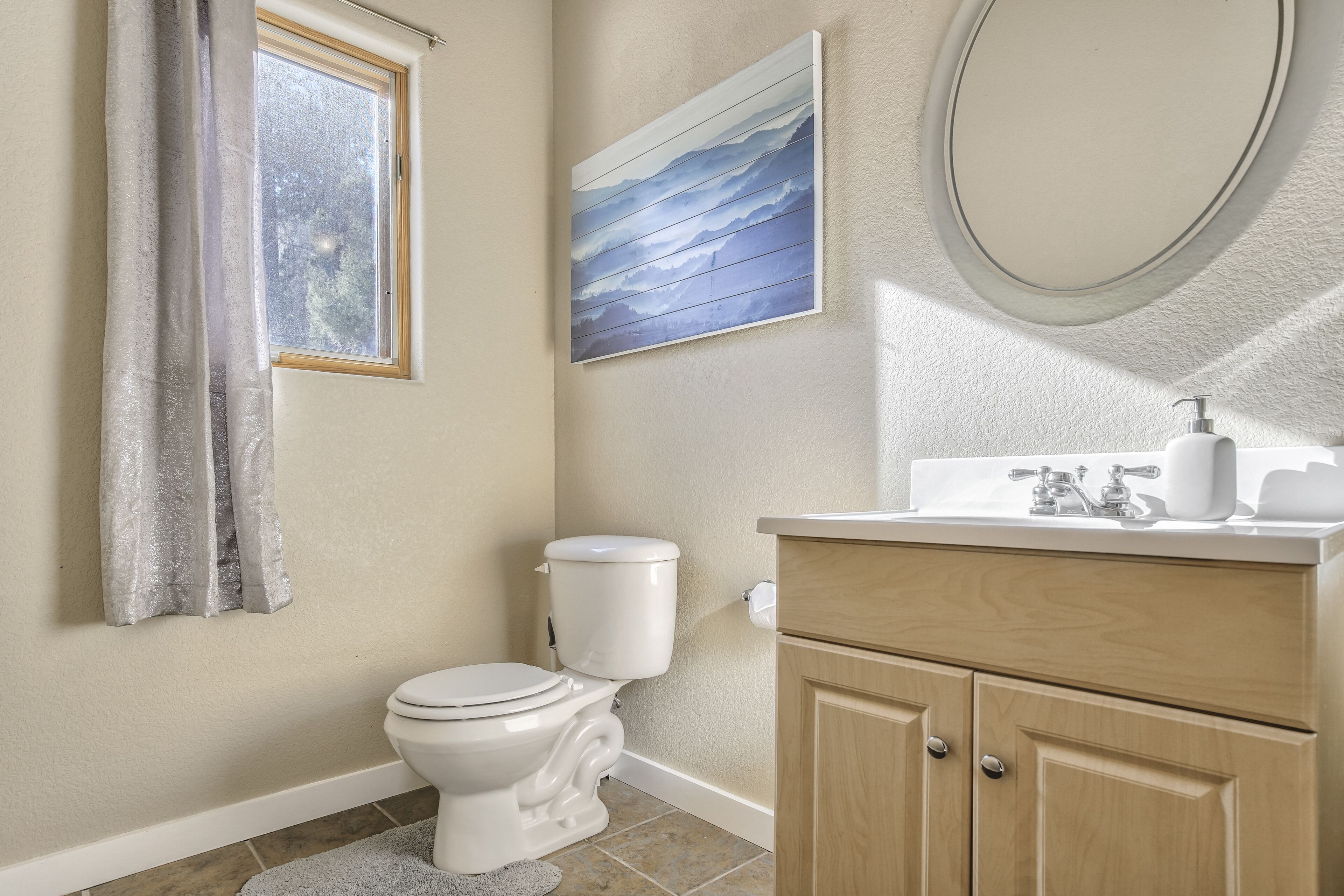 A bathroom with a toilet, a wooden cabinet, a sink with a mirror, and a window with a curtain.