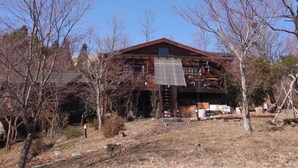 A rustic wooden cabin with a sloped roof, surrounded by leafless trees and a dry grassy area.