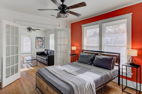 A bedroom with a bed, a ceiling fan, a window with blinds, a nightstand, and a wall-mounted lamp.