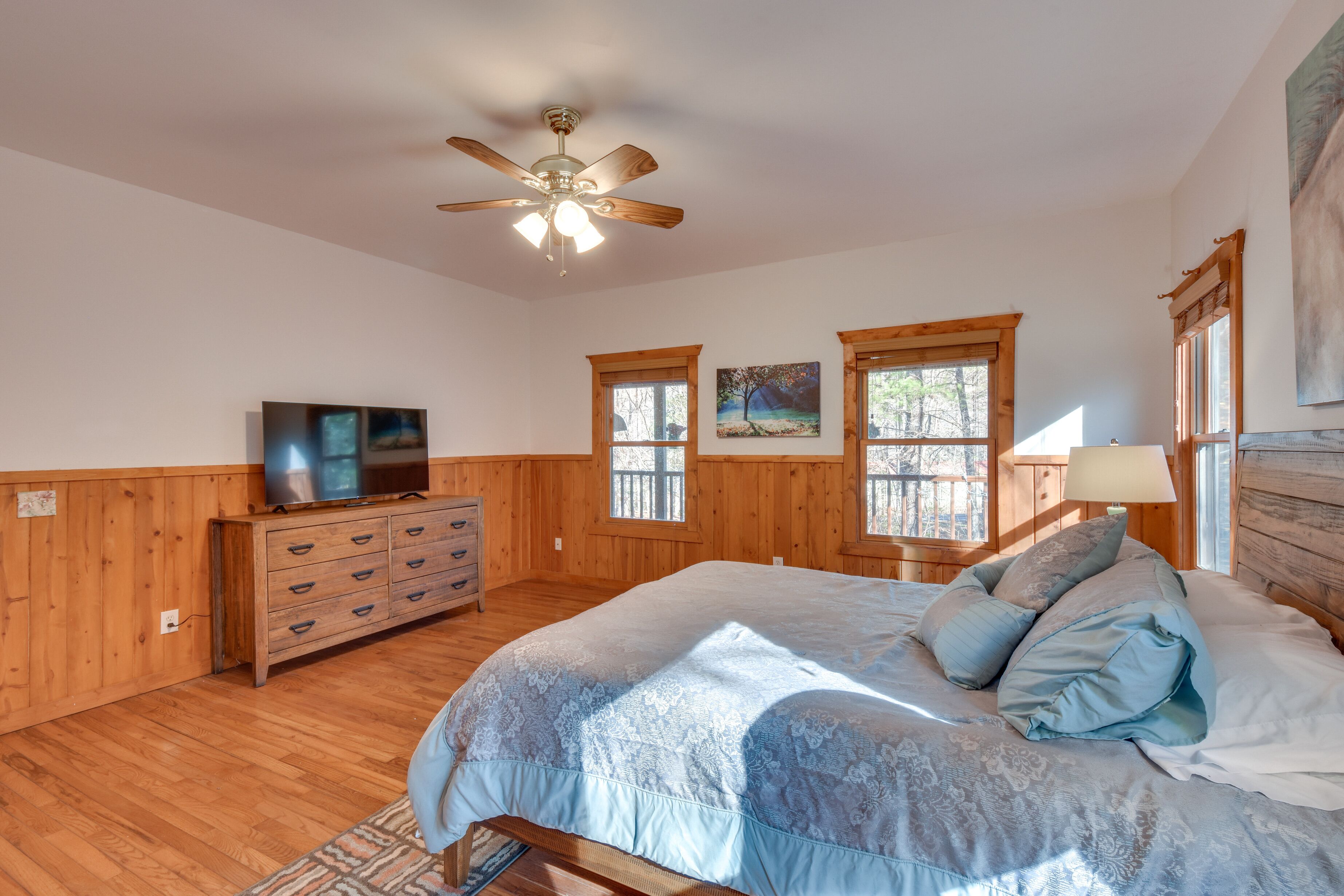 A bedroom with a bed, wooden dresser, television, windows, and a ceiling fan.