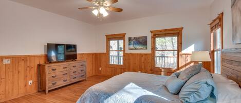 A bedroom with a bed, wooden dresser, television, windows, and a ceiling fan.