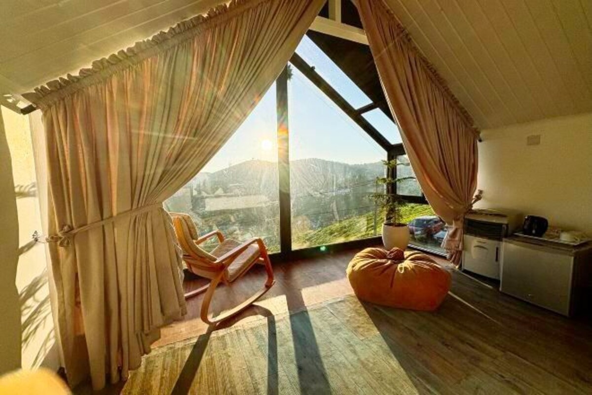 A room with a large window, a wooden floor, a chair, a bean bag, and a kitchenette.