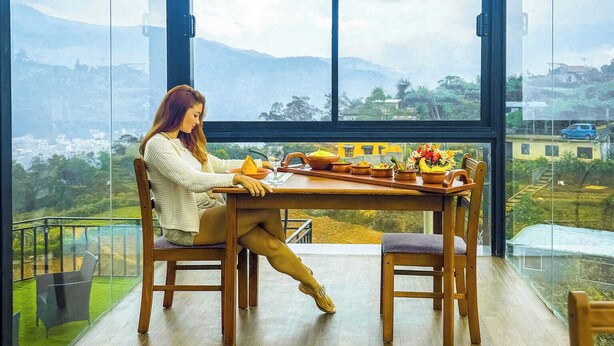 A woman sitting at a wooden table inside a room with large windows, a glass ceiling, and hanging light bulbs. - The Blackberry Cottage (Nuwara Eliya)