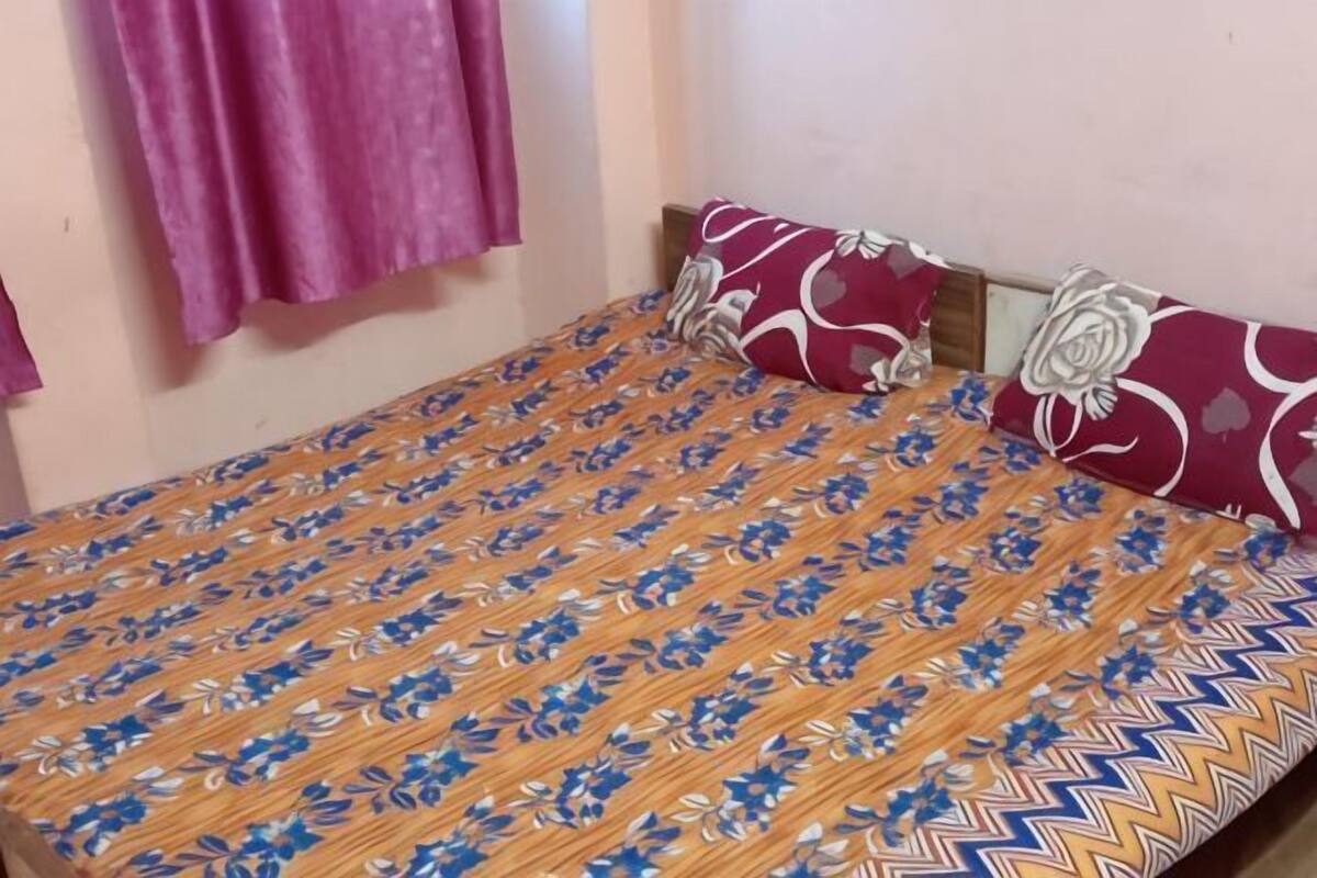 A single bed with a patterned bedspread, a pillow with a floral design, and a window with pink curtains.
