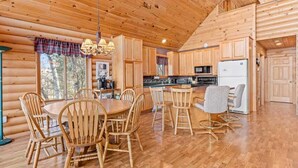 A log cabin interior with a wooden dining table and chairs, a kitchen with wooden cabinets, and a large window. - Hill Road Chalet on Benoit Lake in Spooner WI (Spooner)