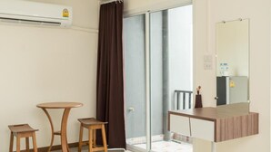 A hotel room with a sliding glass door to a balcony, a small wooden table, a chair, a mirror, and an air conditioning unit. - My Room Nakhonpathom (Nakhon Pathom)