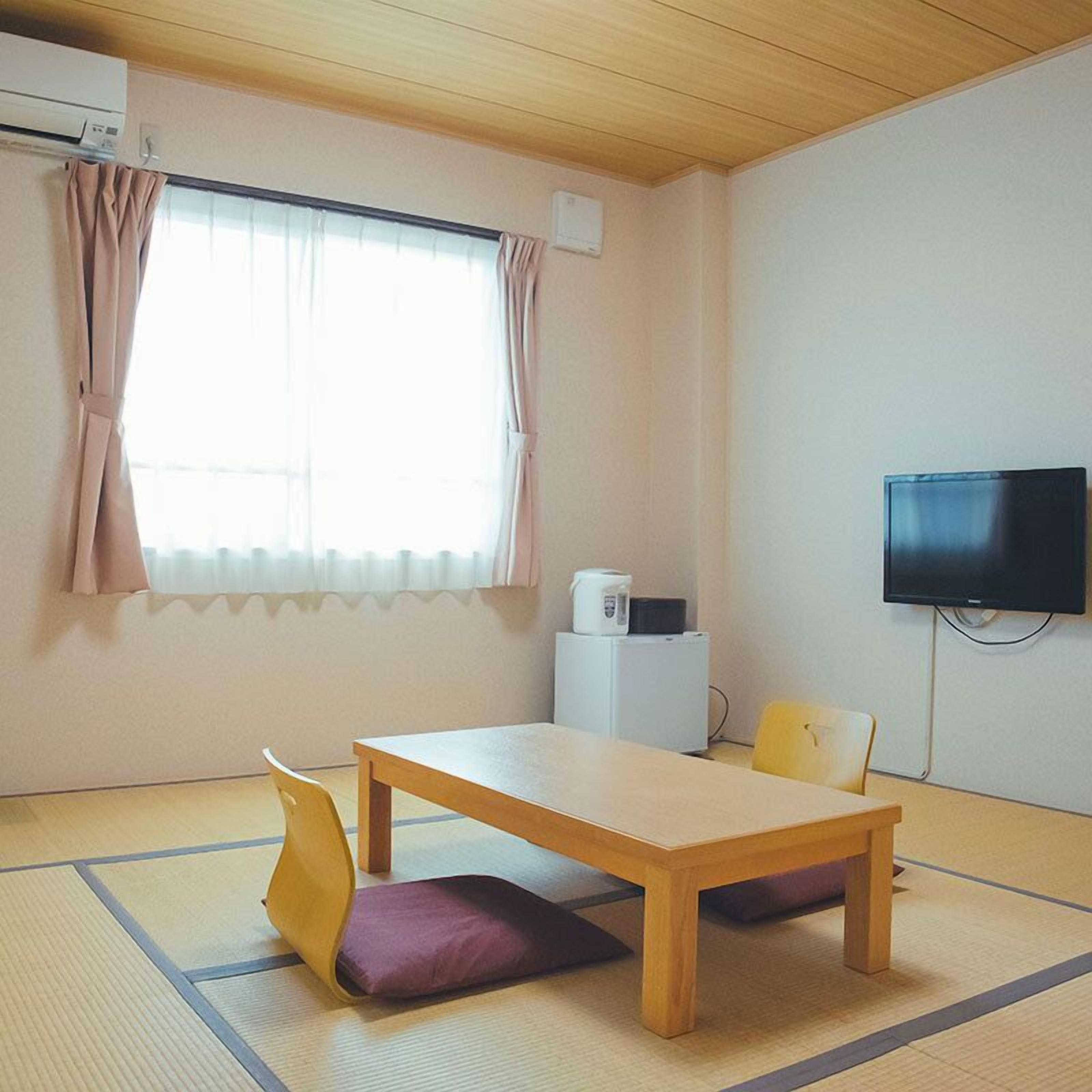 Una habitación con suelo de tatami, una mesa de madera, una silla amarilla, un cojín morado, un refrigerador blanco, un televisor y una ventana con cortinas.