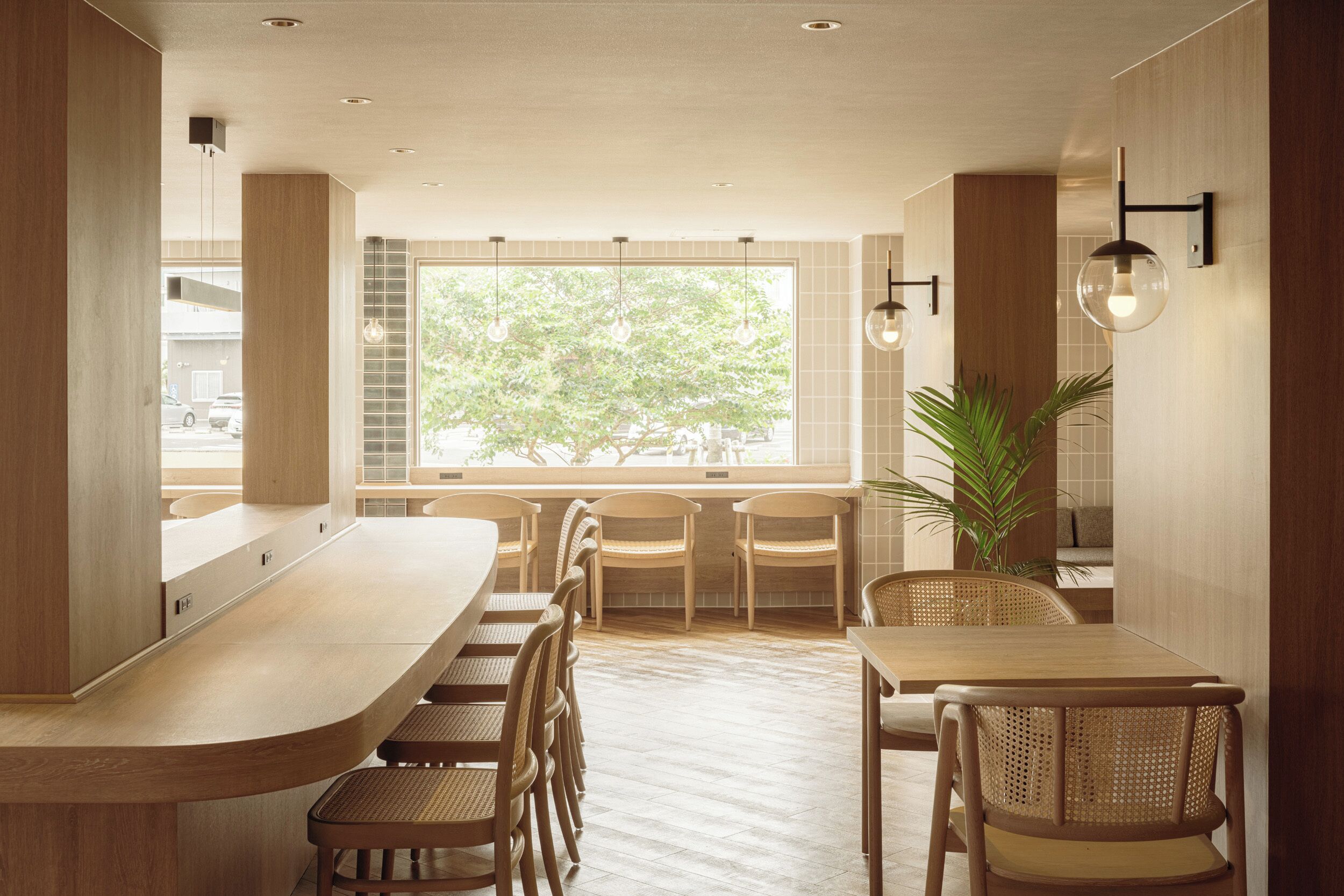A modern restaurant interior with a long counter, wooden chairs, and a large window offering a view of trees.