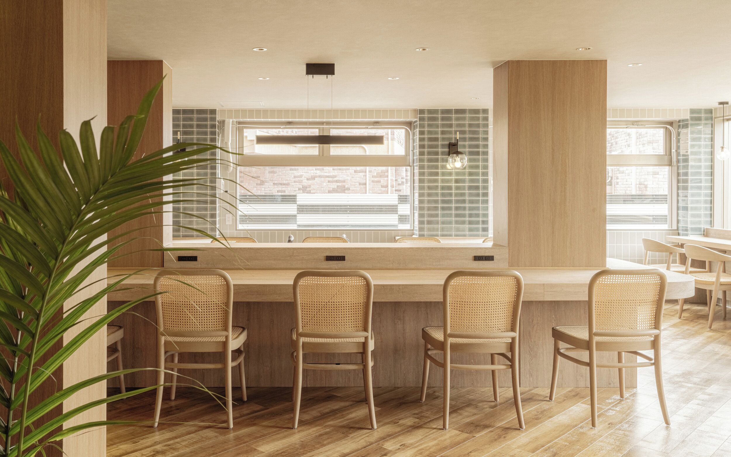 Een moderne bar met een houten toonbank, drie rieten stoelen en een groot raam met zonwering.