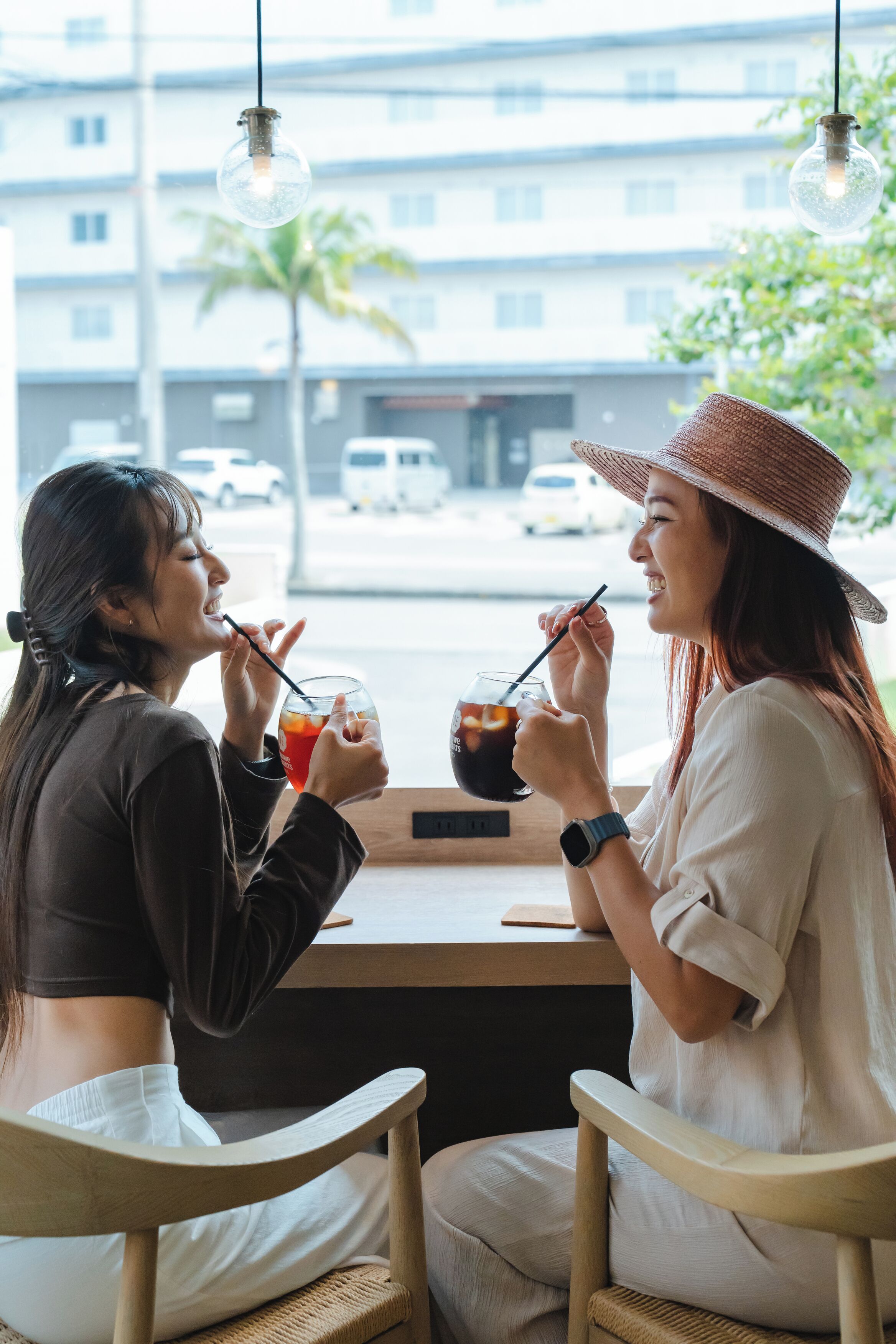 Two people sitting at a table with drinks, in a room with a view of buildings and a palm tree outside.