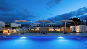 An outdoor swimming pool area with multiple umbrellas, seating, and a view of the cityscape at dusk. - Nest Hotel Ishigaki Maesato Beach (Ishigaki)