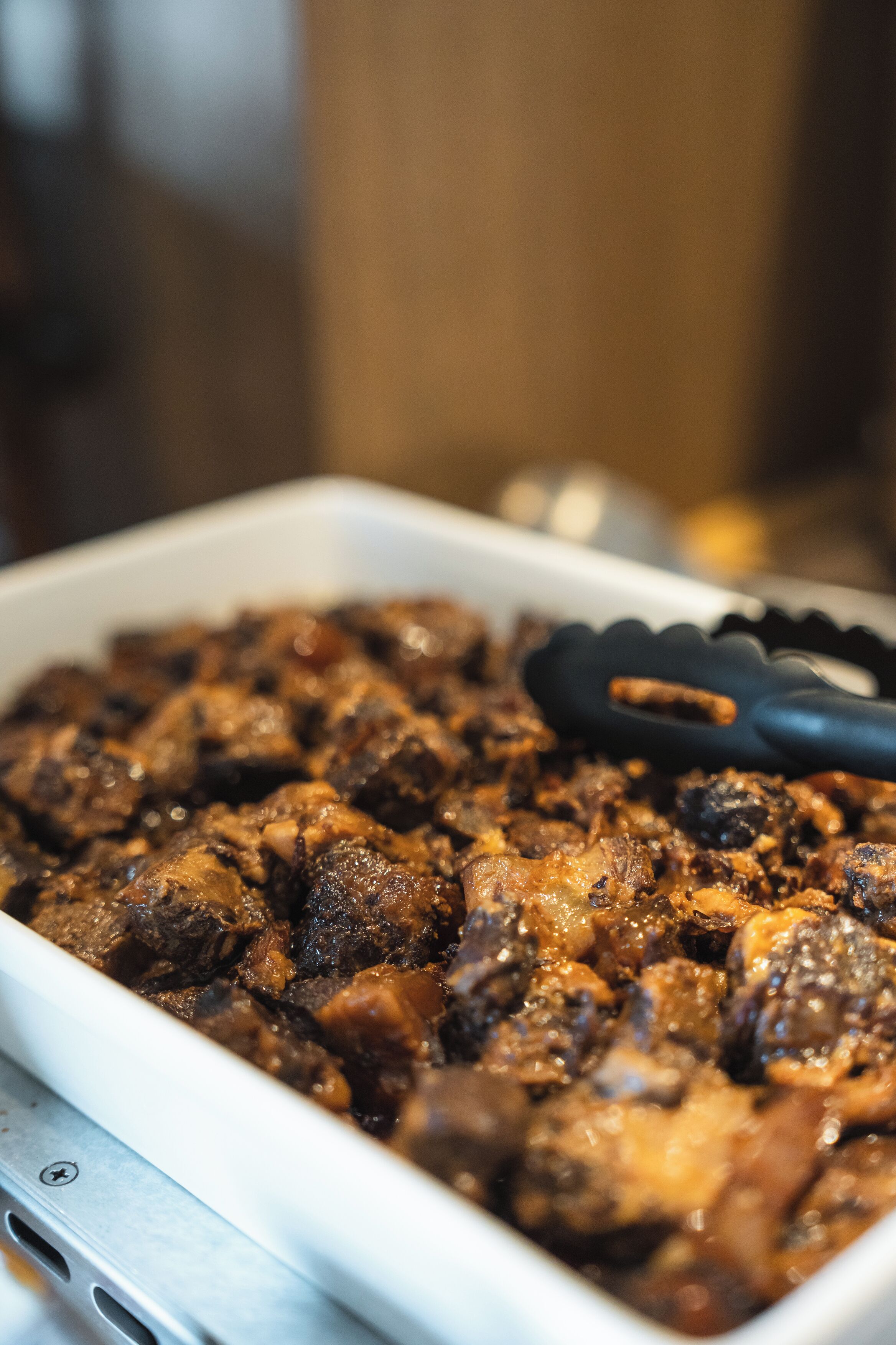 A white rectangular dish containing a dark brown, chunky food item, possibly a stew or casserole, with a pair of black tongs resting beside it.