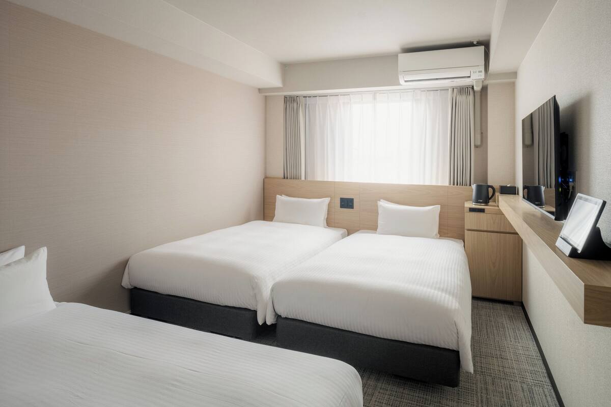Ein Hotelzimmer mit zwei Betten, einem Schreibtisch mit Stuhl, einem Flachbildfernseher und einer Klimaanlage.