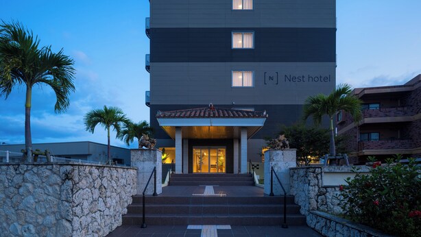A multi-story hotel with a modern facade, a tiled entrance, and a stone wall with palm trees. - Nest Hotel Ishigaki Maesato Beach (Ishigaki)