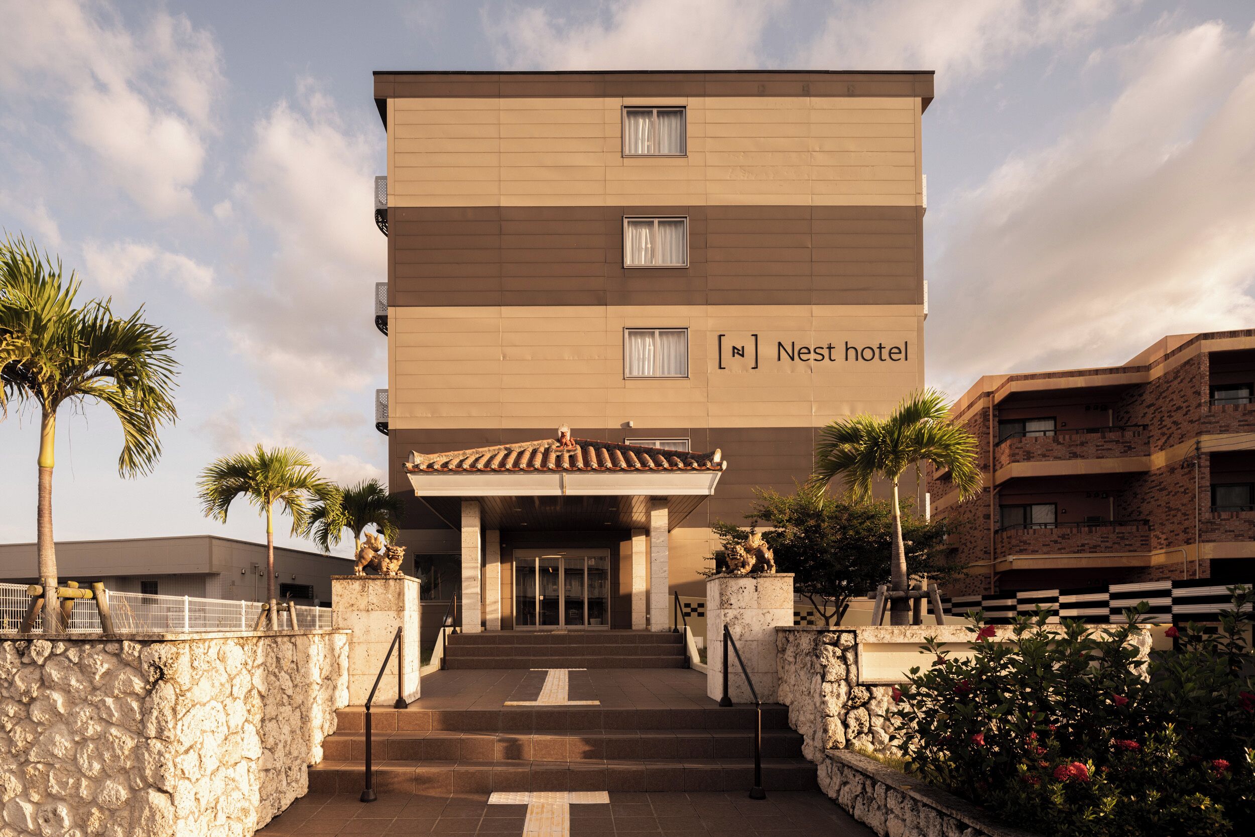 Un hotel a più piani con facciata in pietra, un ingresso centrale con gradini e palme sullo sfondo.