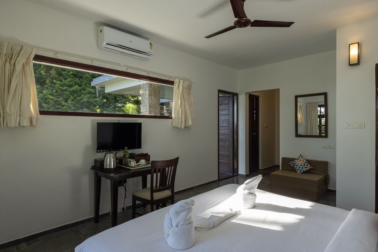 Quarto de hotel com cama, escrivaninha com televisão, poltrona, sofá e ventoinha no teto.