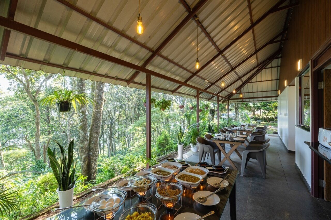 Un área de comedor techada con vista a un paisaje boscoso, mesas dispuestas con comida y plantas colgantes.
