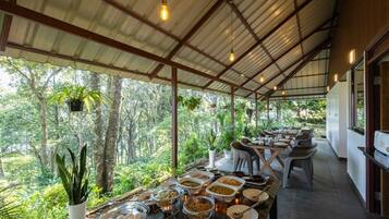 Un área de comedor techada con vista a un paisaje boscoso, mesas dispuestas con comida y plantas colgantes.