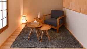 Ein kleiner, gemütlicher Raum mit Holzboden, einem grauen Teppich, einem runden Holztisch, einem blauen Stuhl, einer hängenden Lampe und einem gerahmten Bild an der Wand.