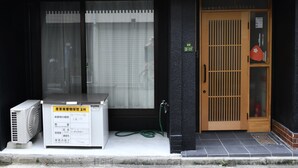 Eine Gebäudefassade mit vertikalen Holzlamellen, eine Tür mit Gittermuster und eine kleine Klimaanlage am Boden.