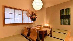 Ein traditioneller japanischer Raum mit Tatami-Bodenbelag, einem niedrigen Holztisch mit Stühlen, einer hängenden Papierlaterne, einem Fenster mit Shoji-Schirm und einem gerahmten Kunstwerk an der Wand.