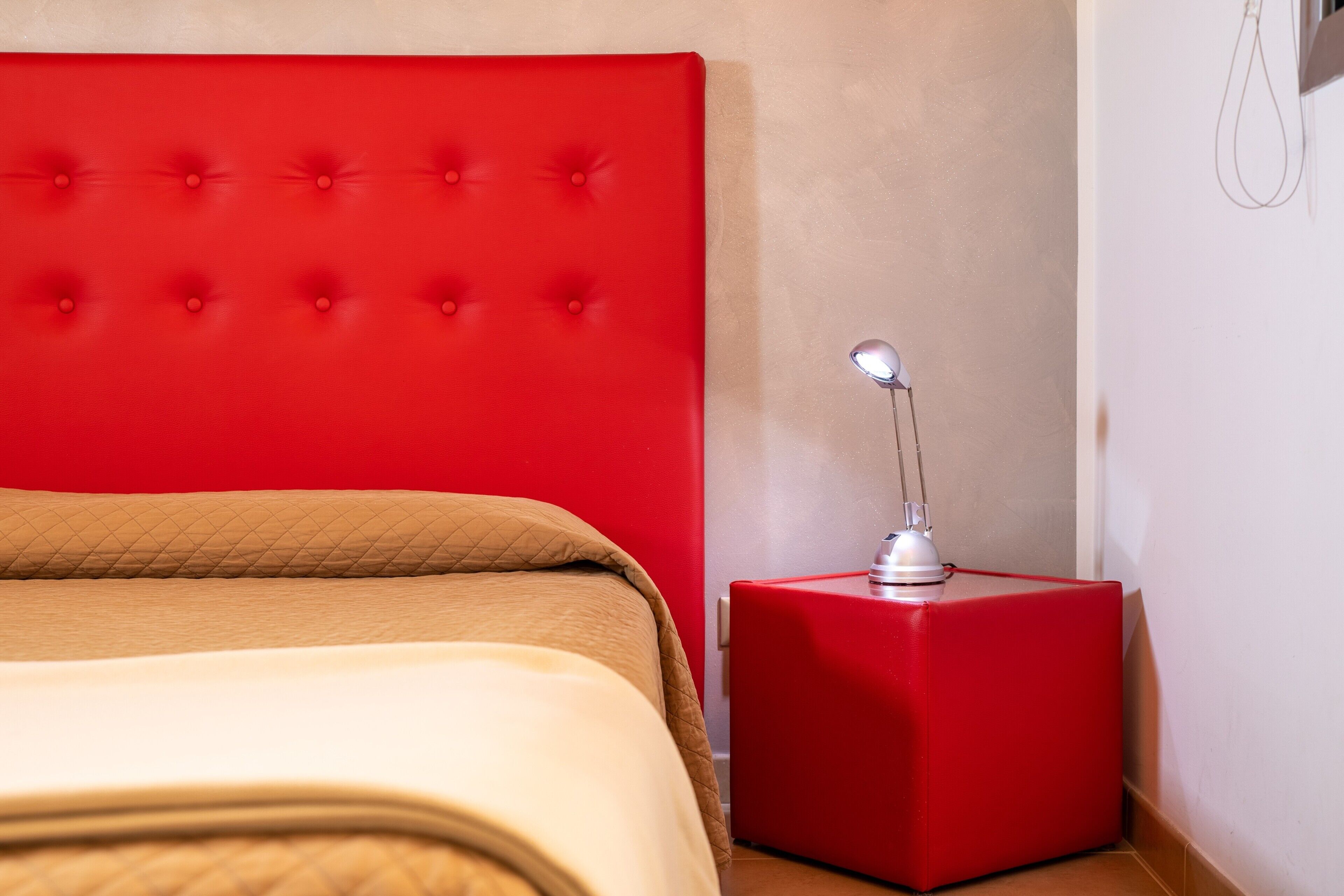 Een bed met een rood hoofdeinde, een nachtkastje in rood met een lamp, en een beige deken.