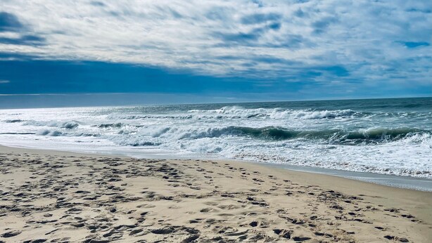 A beach with footprints leading to the water, waves crashing, and a partly cloudy sky.