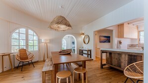 A spacious kitchen with a wooden table, two stools, a hanging light, and a view through an arched window.