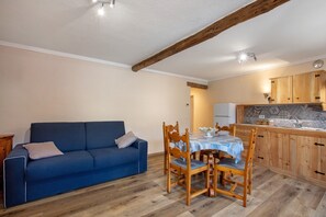 Une pièce avec un canapé bleu, une table à manger en bois et des chaises, une cuisine avec des meubles en bois et un plafond avec des poutres apparentes.