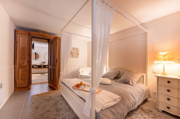 A bedroom with a four-poster bed, a bedside table with a lamp, a wooden wardrobe, and a bathroom visible through an open door.