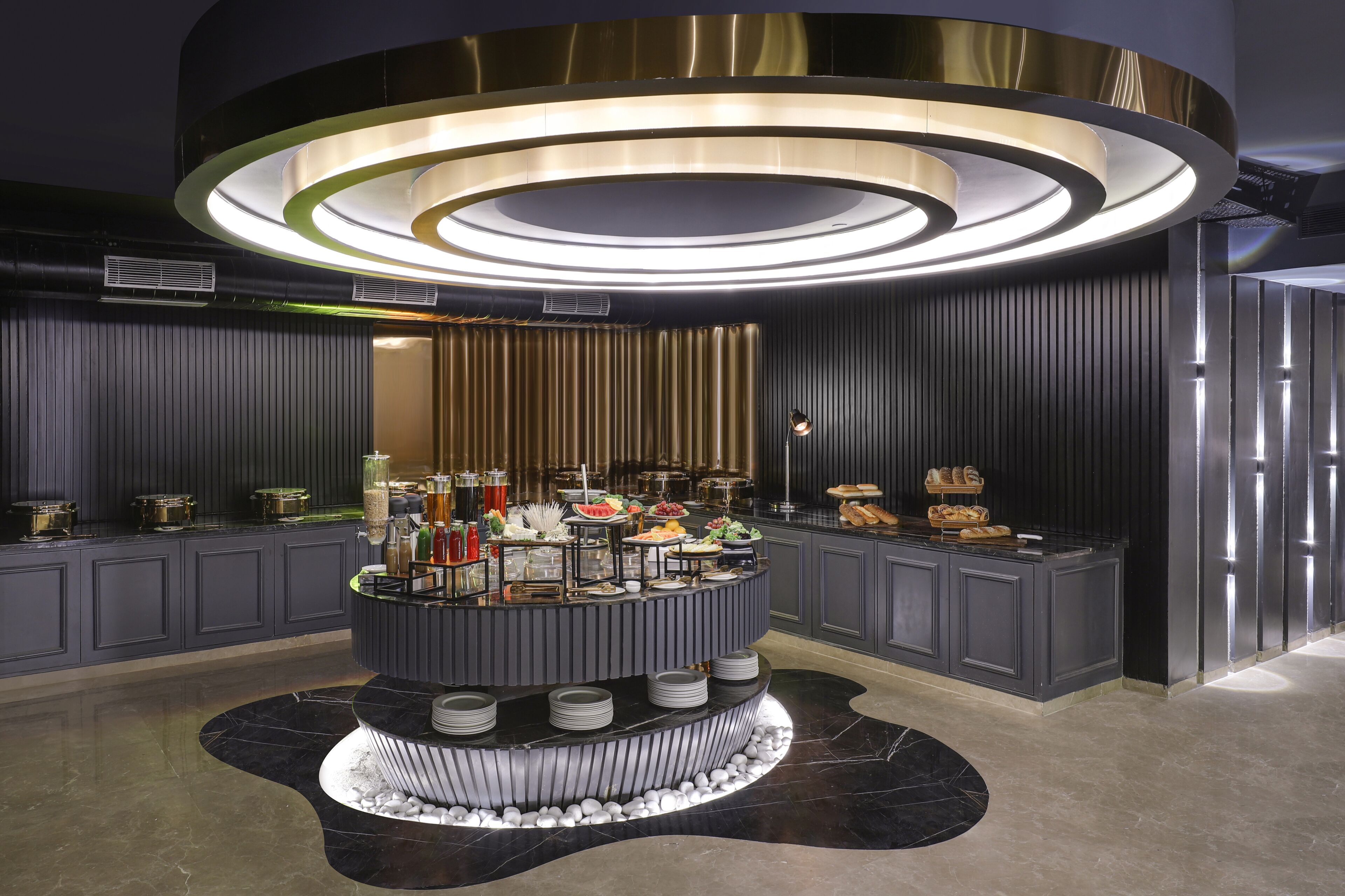 A modern buffet setup with a central island, surrounded by dining tables and a decorative ceiling light fixture.
