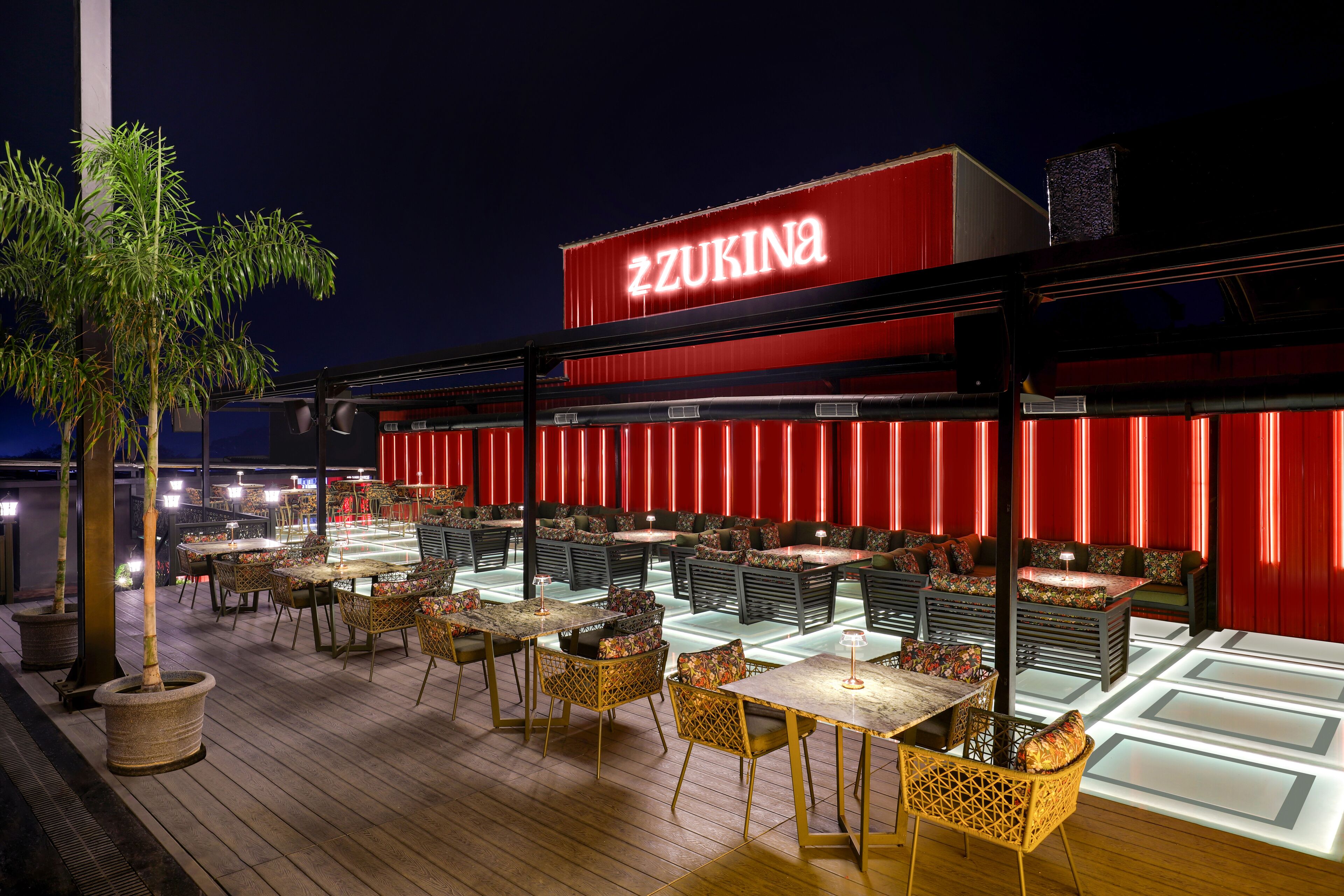 An outdoor dining area with tables and chairs, a red building with illuminated signage, and potted plants.