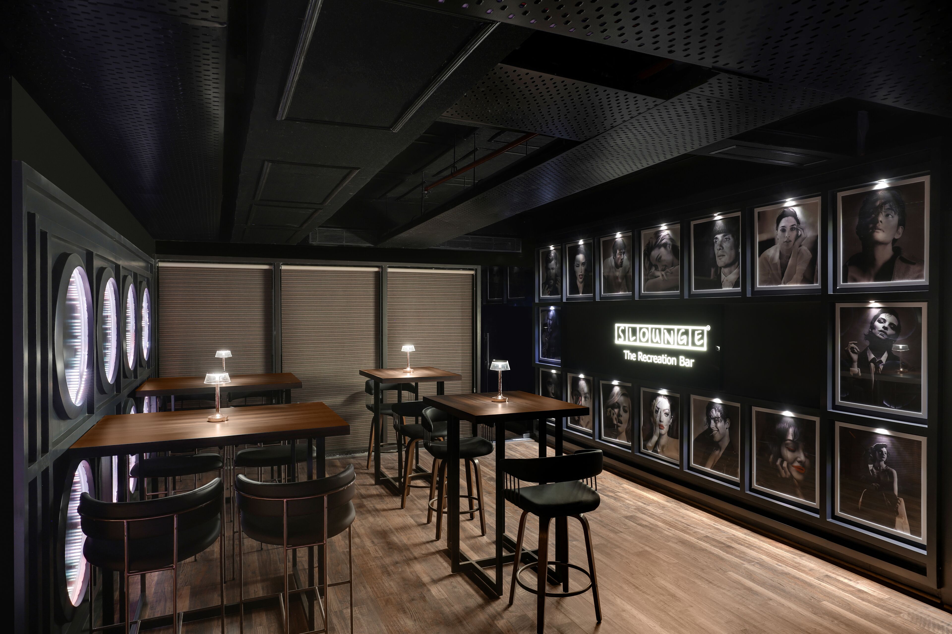A dimly lit bar area with wooden tables and black bar stools, featuring framed portraits on the wall and a neon sign reading "SLOUNGE The Recreation Bar".