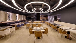 Ein modernes Restaurant mit einer zentralen, runden Lichtinstallation, mehreren Esstischen und zeitgenössischen Stühlen.