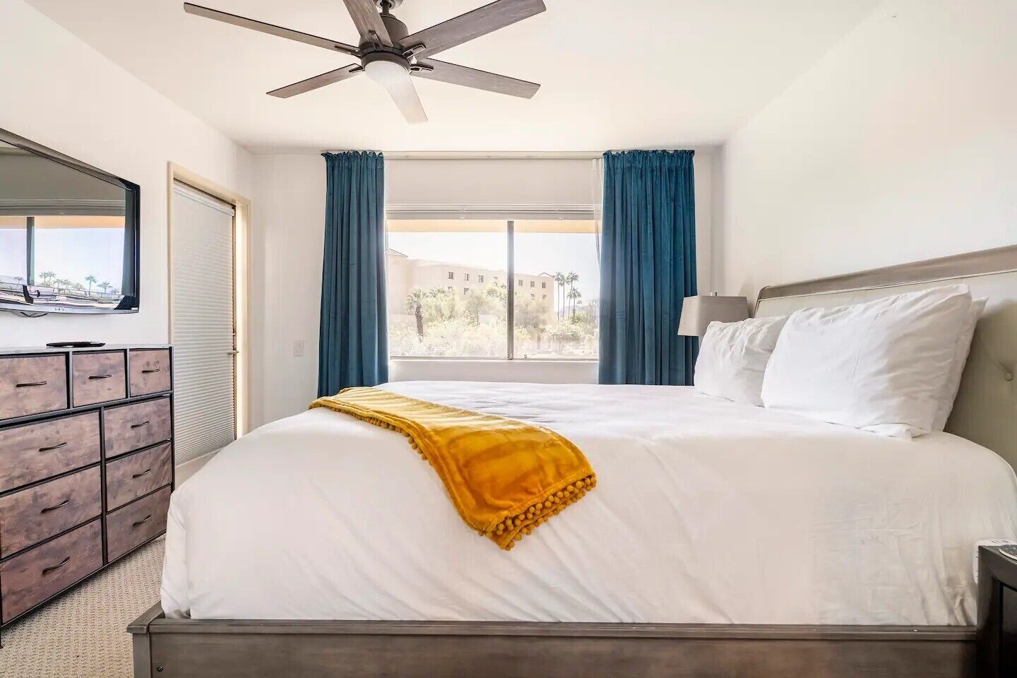 Une chambre d’hôtel avec un lit, un ventilateur de plafond, une commode et une fenêtre avec des rideaux.