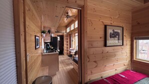 A wooden cabin interior with a kitchen area, a dining table, and a bed with a red blanket. - Ptarmigan Tiny Home (#10) at The Woolly Bugger Inn (Estes Park)