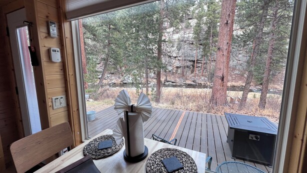 A wooden interior with a large window overlooking a forested area, a table with a notebook, a decorative vase, and a grill outside. - Ptarmigan Tiny Home (#10) at The Woolly Bugger Inn (Estes Park)