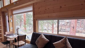 A cozy cabin interior with a sofa, a desk, and large windows offering a view of a forested area. - Ptarmigan Tiny Home (#10) at The Woolly Bugger Inn (Estes Park)