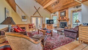 A spacious living room with a high ceiling, wooden beams, a fireplace, a television, and a large rug. - Cimmaron Sky by Velo (Angel Fire)