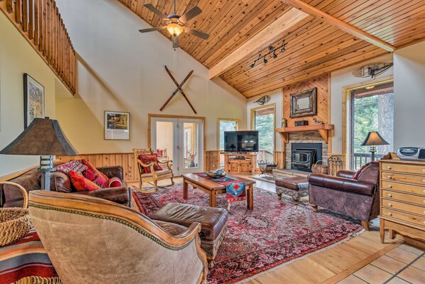 A spacious living room with a high ceiling, wooden beams, a fireplace, a television, and a large window offering a view of the outdoors.