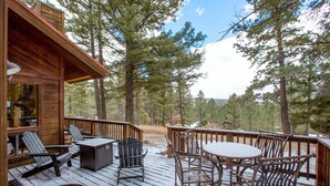 Una terraza de madera con mobiliario exterior, incluyendo sillas y una mesa, con vista a un área boscosa.