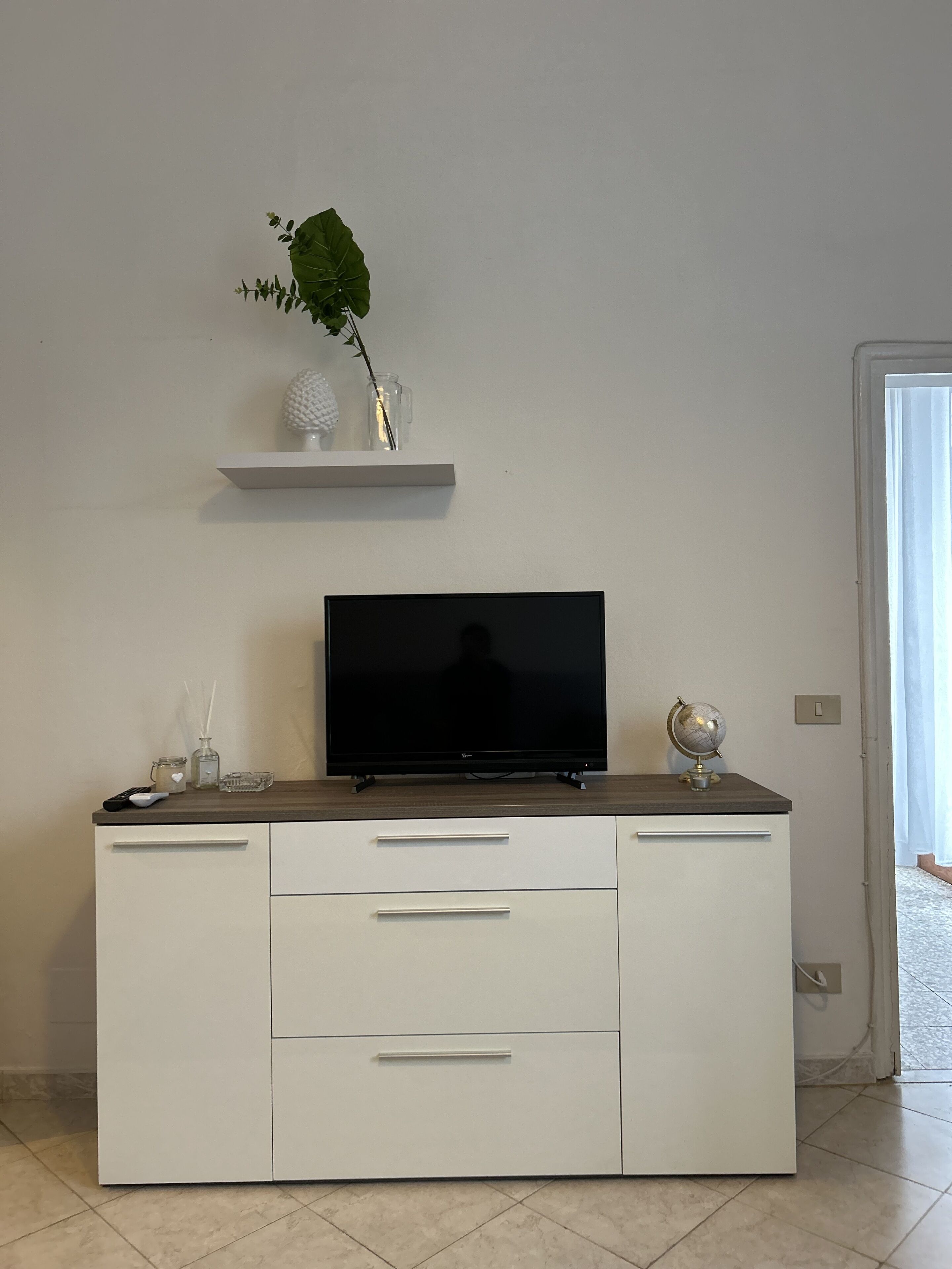 A modern living room with a flat-screen TV on a wooden cabinet, a small shelf with decorative items, and a door leading to another room.
