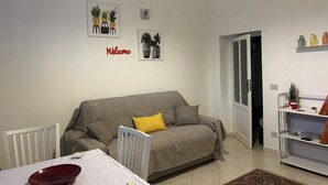 Un salon avec un canapé gris, un fauteuil blanc, une table sur laquelle se trouve un bol rouge, et un panneau « Bienvenue » accroché au mur.
