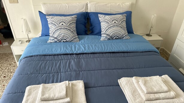 Un lit bien fait, recouvert d’une couverture bleue, avec deux oreillers blancs à motifs bleus et deux serviettes blanches pliées posées sur le lit.