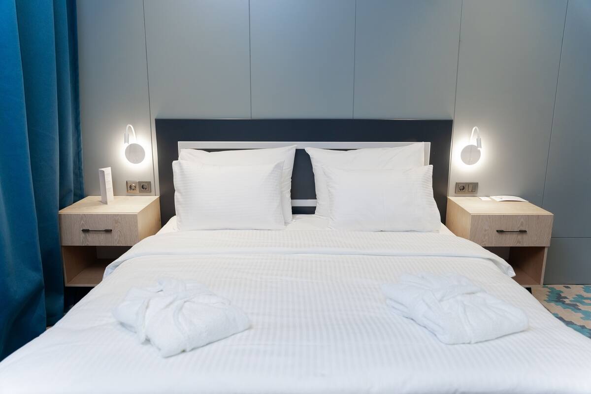 A neatly made bed with white linens, two bedside tables with illuminated wall sconces, and a blue curtain on the left.