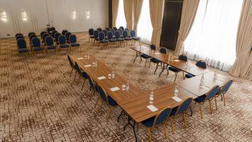 Un salón de conferencias con varias mesas y sillas, piso alfombrado y ventanas grandes con cortinas.