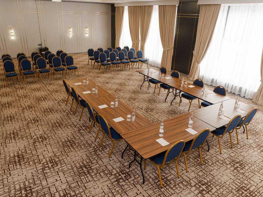 Un salón de conferencias con varias mesas y sillas, piso alfombrado y ventanas grandes con cortinas.