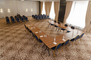 Un salón de conferencias con varias mesas y sillas, piso alfombrado y ventanas grandes con cortinas.