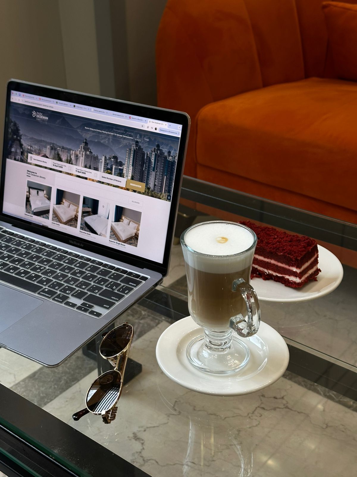A laptop on a table displaying a hotel website, a glass of coffee, and a slice of cake.